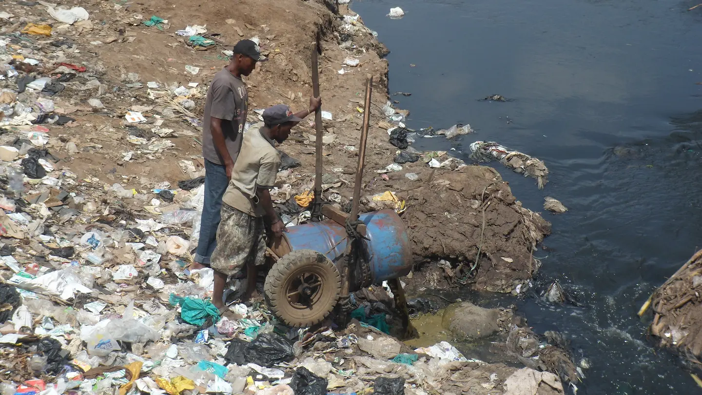 2人の男性が、収集したし尿汚泥を地元の川へ投棄している様子。周囲の地面には多くのごみが散乱している。