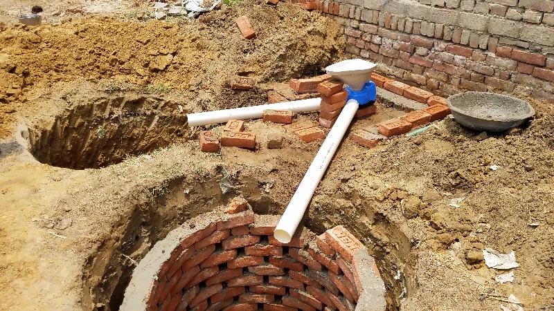 建設中の二槽式簡易水洗トイレ。穴を掘ってレンガで内壁をライニングしたピットが2つあり、白い塩ビパイプとスクワット式便器が仮設で配置されている。周囲は土とレンガで施工途中の様子が見える。