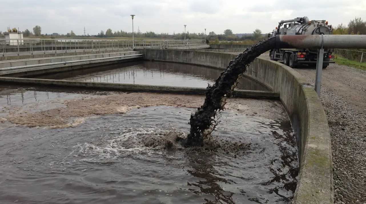 汚泥濃縮池へ、太い排水管から黒く濁った汚泥が勢いよく投入されている実写風の写真。投入された汚泥は水面で泡立ちながら、重力によって池の底部へと沈殿していく。