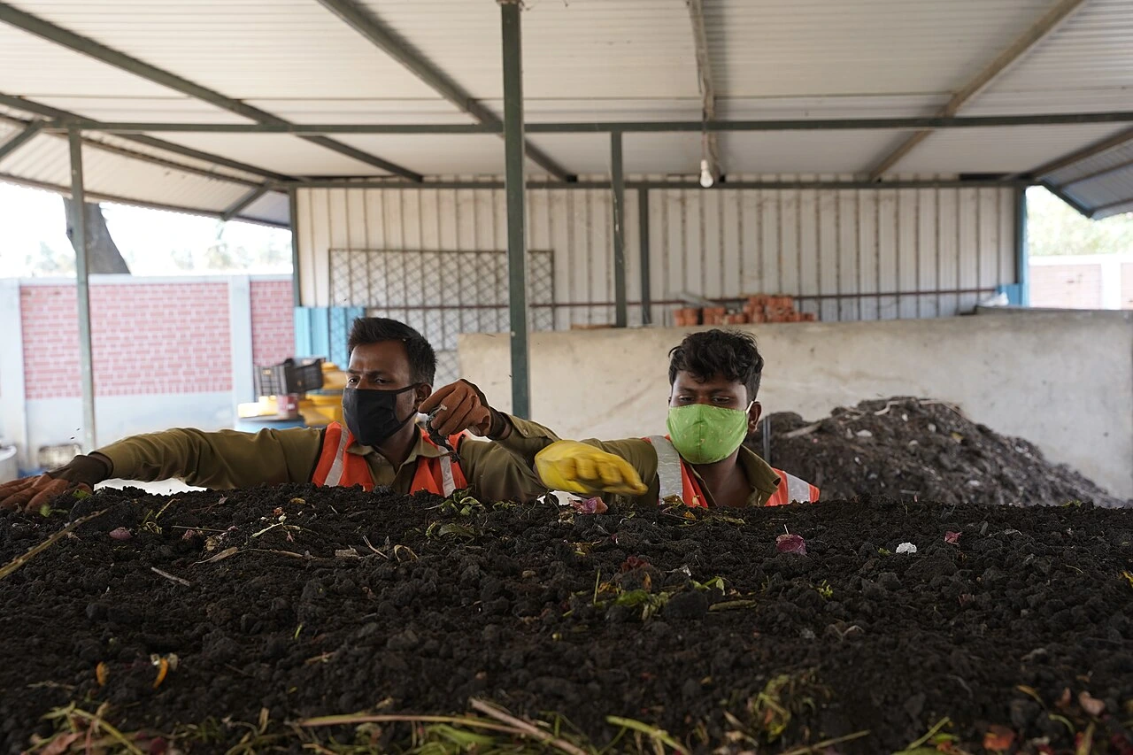 インドのベンガルール（デバナハリ町）における、し尿汚泥と都市生ごみの混合コンポスト化（Windrow Co-composting）の様子を捉えた写真です。 屋根付きの施設内で、オレンジ色の安全ベストとマスクを着用した2人の作業員が、手前にある黒々とした堆肥（コンポスト）の山を素手やヘラを使って丁寧に手入れしています。奥にはさらに高く積み上げられた堆肥の山が見え、汚泥と生ごみを混ぜ合わせて資源化する、大規模なリサイクルプロセスの現場を伝えています。