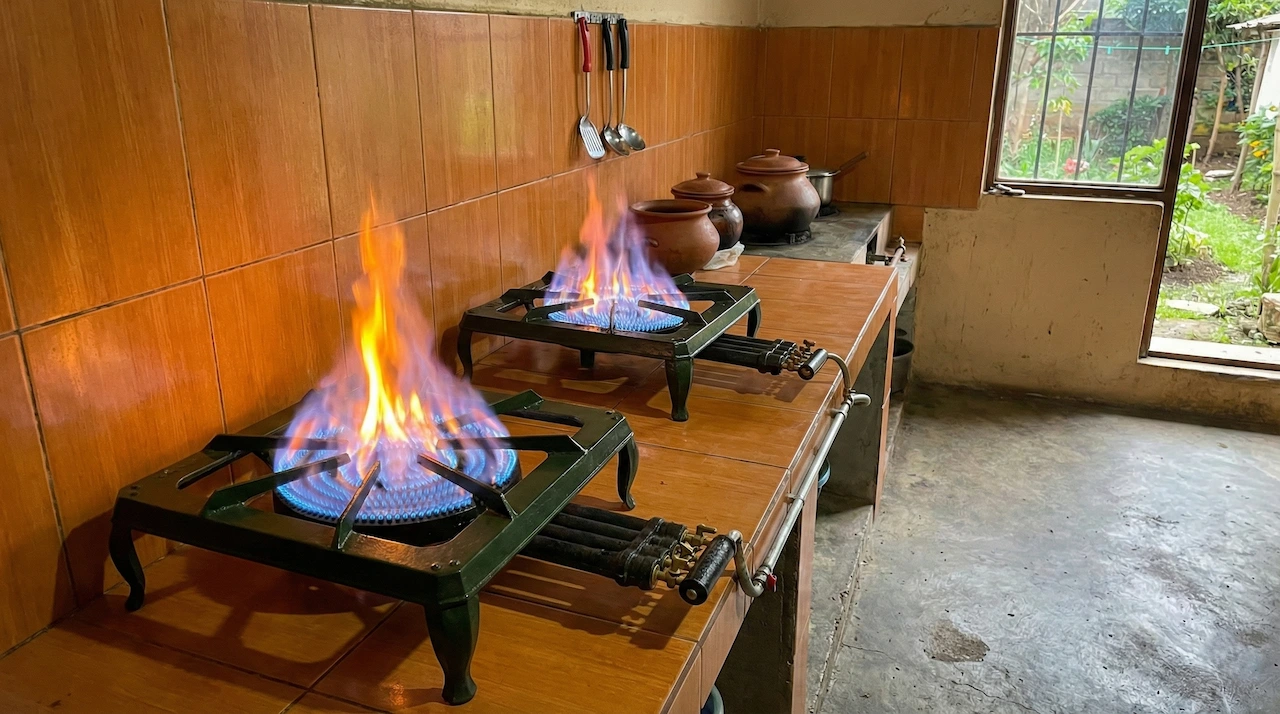 オレンジ色のタイル張りのキッチンカウンターに設置された2つのバイオガスコンロが、青とオレンジの炎を上げて燃焼している様子を捉えた写真。コンロは緑色の金属製で、ガス管が接続されている。背景には壁に掛かった調理器具や複数の土鍋があり、窓の外には緑豊かな庭が見える。