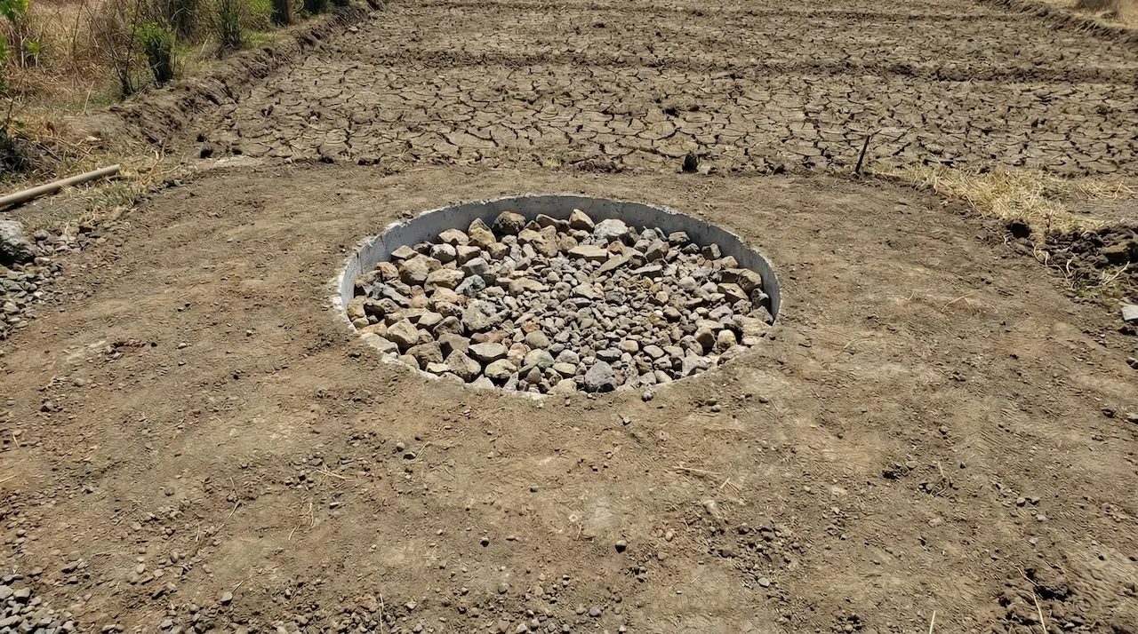 乾いた地面に作られた、蓋のない完成した浸透ピット（soak-pit）の写真です。円形の穴の内部には砂利が上端までぎっしりと詰め込まれており、排水がこれらの石の間を通って地中に浸透していく構造がよく分かります。ピットの周囲は乾燥してひび割れた茶色の土壌で、建設作業の跡が見られます。